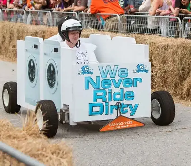 A person wearing a helmet drives a soapbox racer decorated like laundry machines with the text