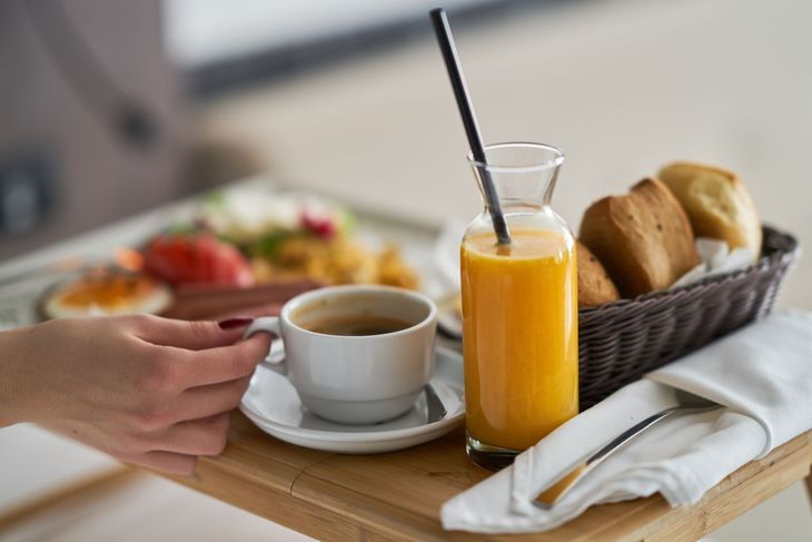 Hand reaching for a cup of coffee beside orange juice, bread basket, and folded napkin on a breakfast table