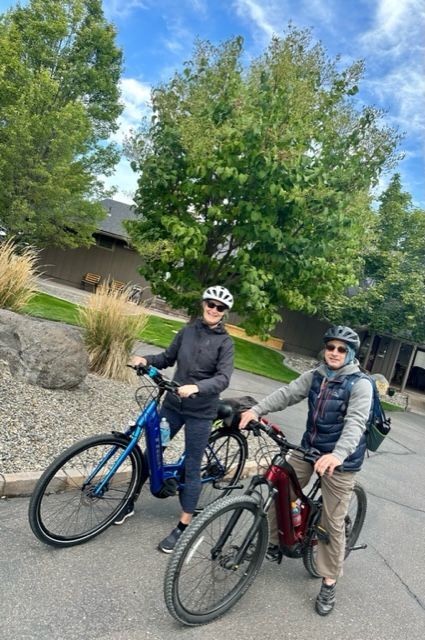Two people wearing helmets on bikes, smiling outdoors near trees.