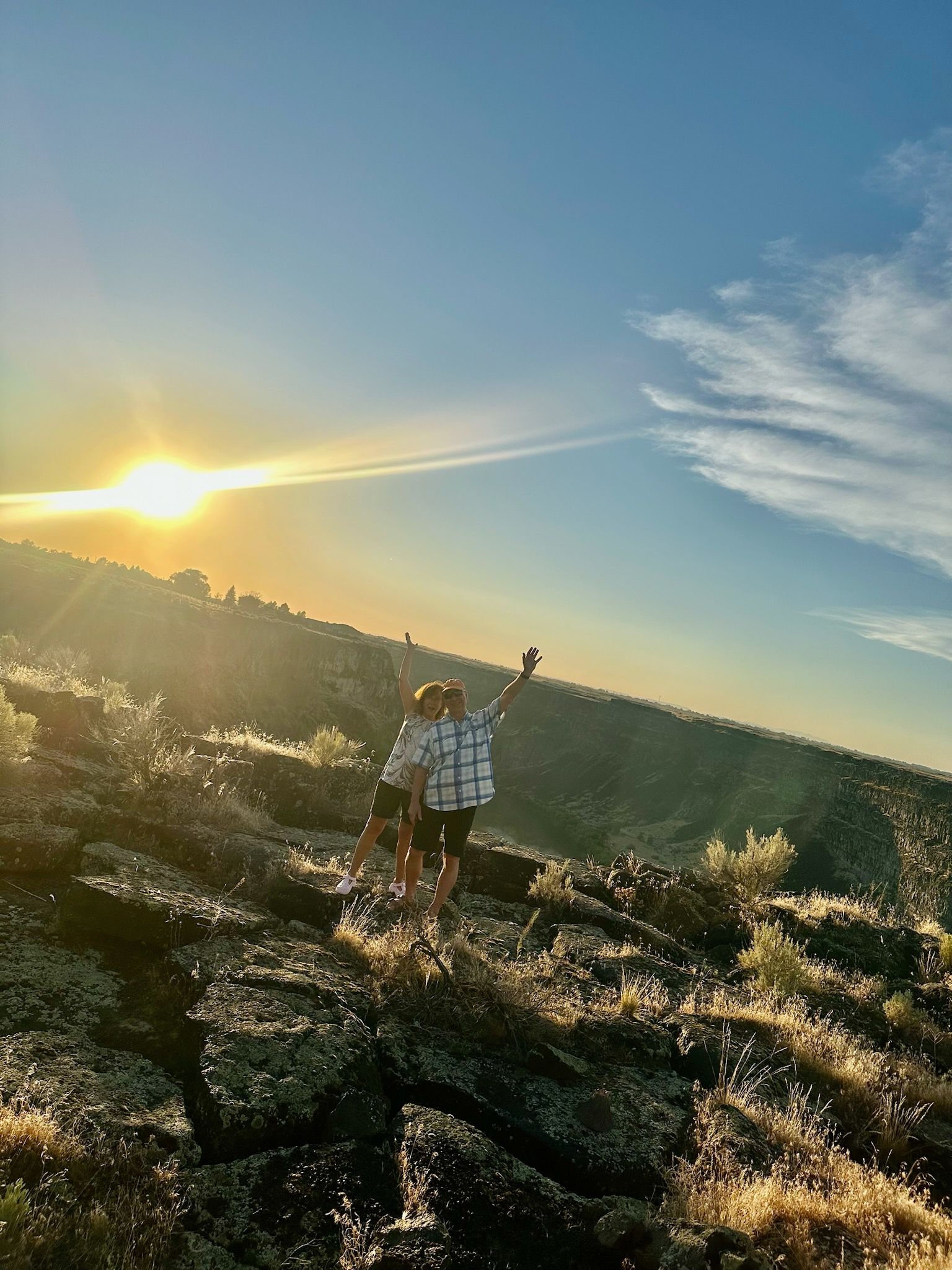 Two people on a rocky hillside at sunset, one with arm raised towards the sun, blue sky overhead.
