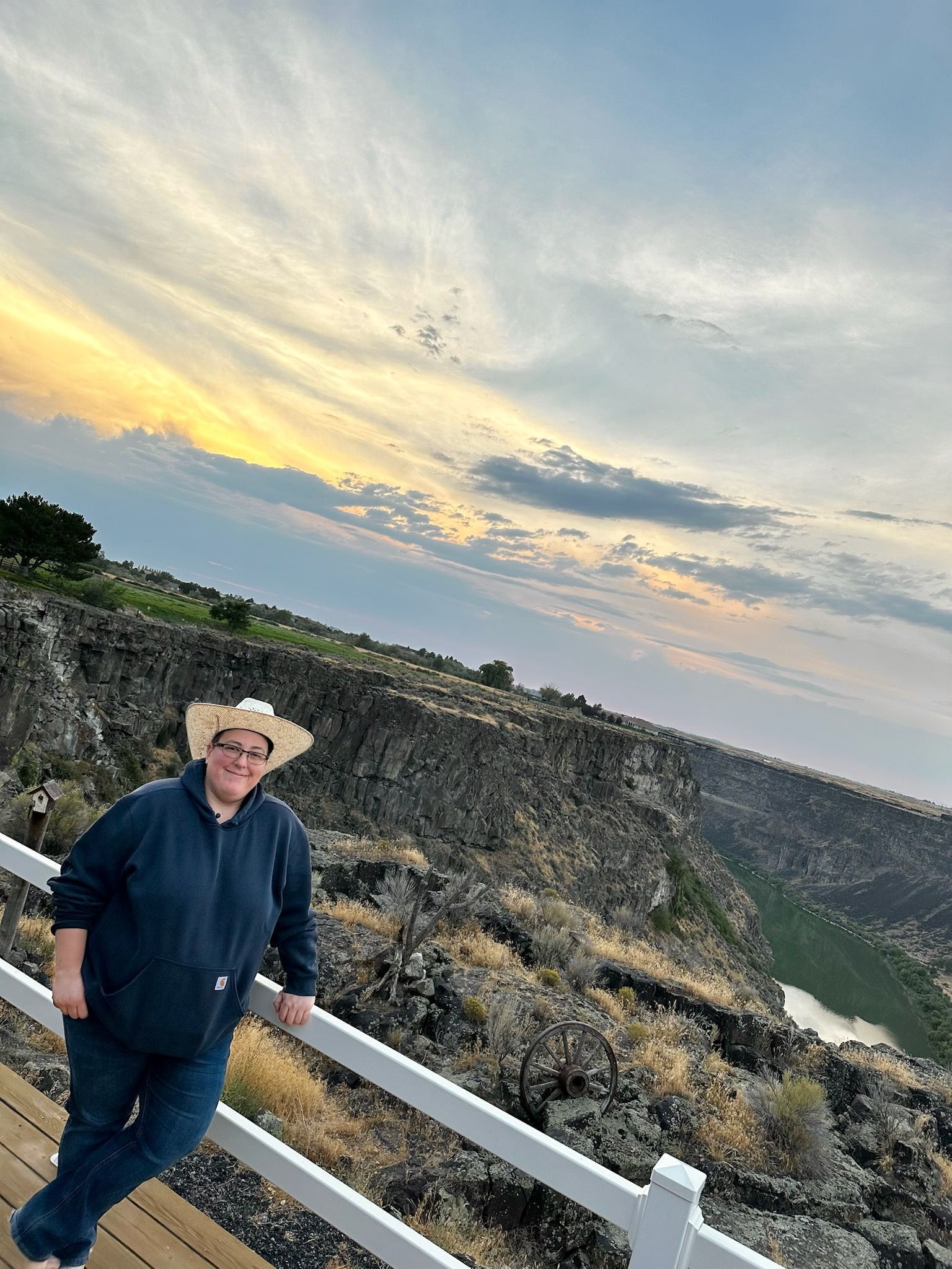 Person in a cowboy hat stands by a white fence at sunset overlooking a rocky canyon.