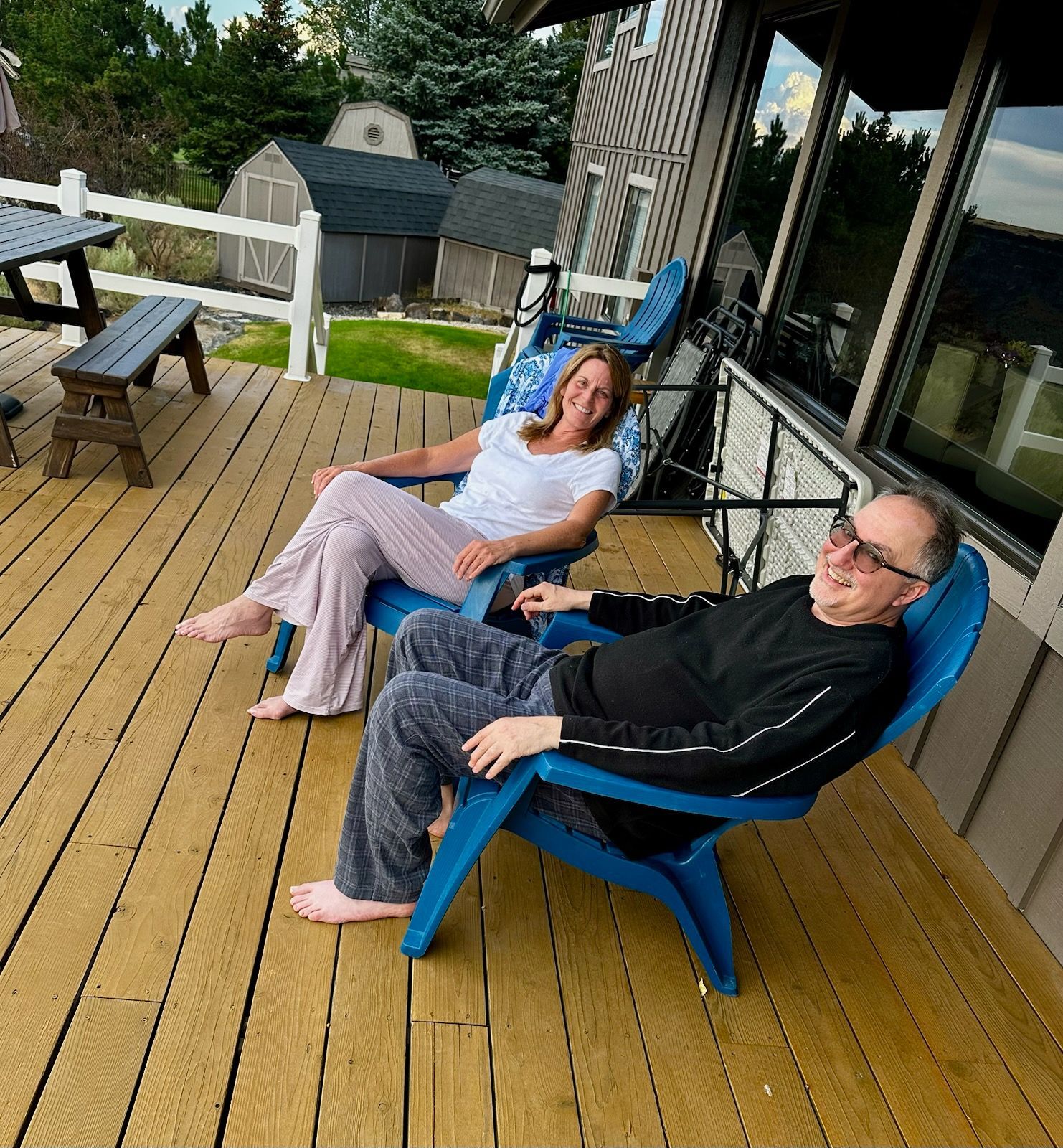 Two people relaxing in blue chairs on a wooden deck beside a house.