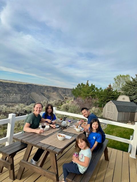 Group seated around picnic table on a deck overlooking a canyon, with a small shed and cloudy sky.