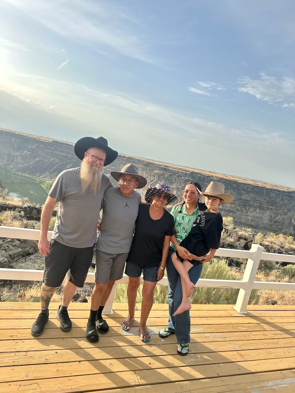 Five people pose outdoors on a wooden platform, wearing hats, smiling, with a canyon in the background.