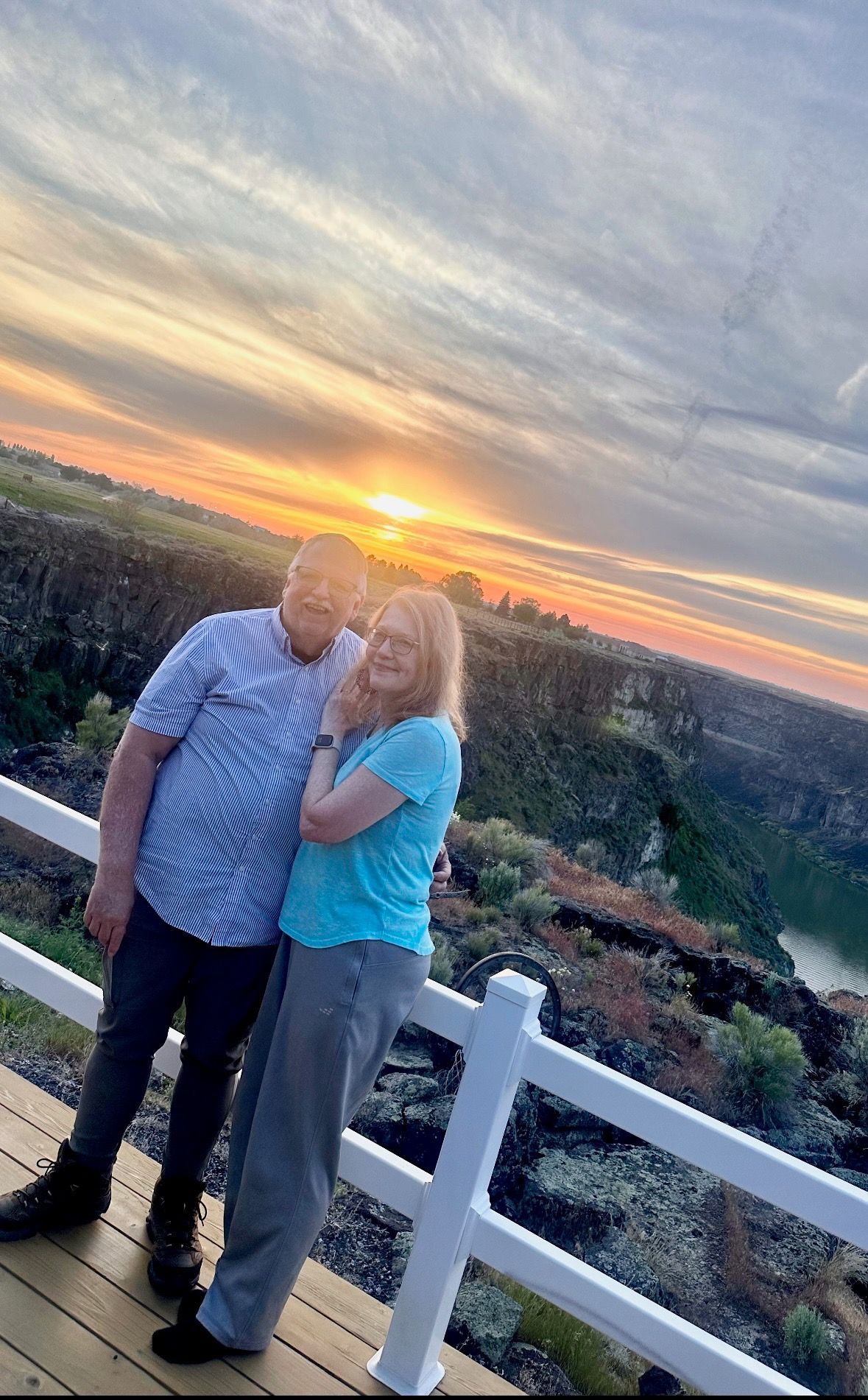 Couple embraces outdoors at sunset, overlooking canyon. Man wears blue shirt, woman in teal.