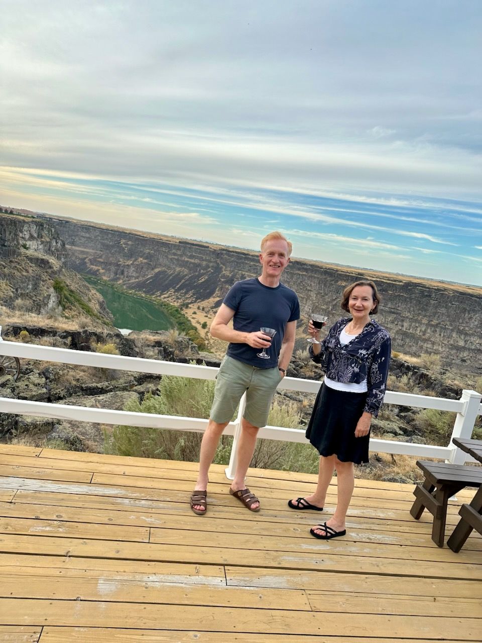 Two people stand on a wooden deck with drinks, overlooking a canyon. Cloudy sky.