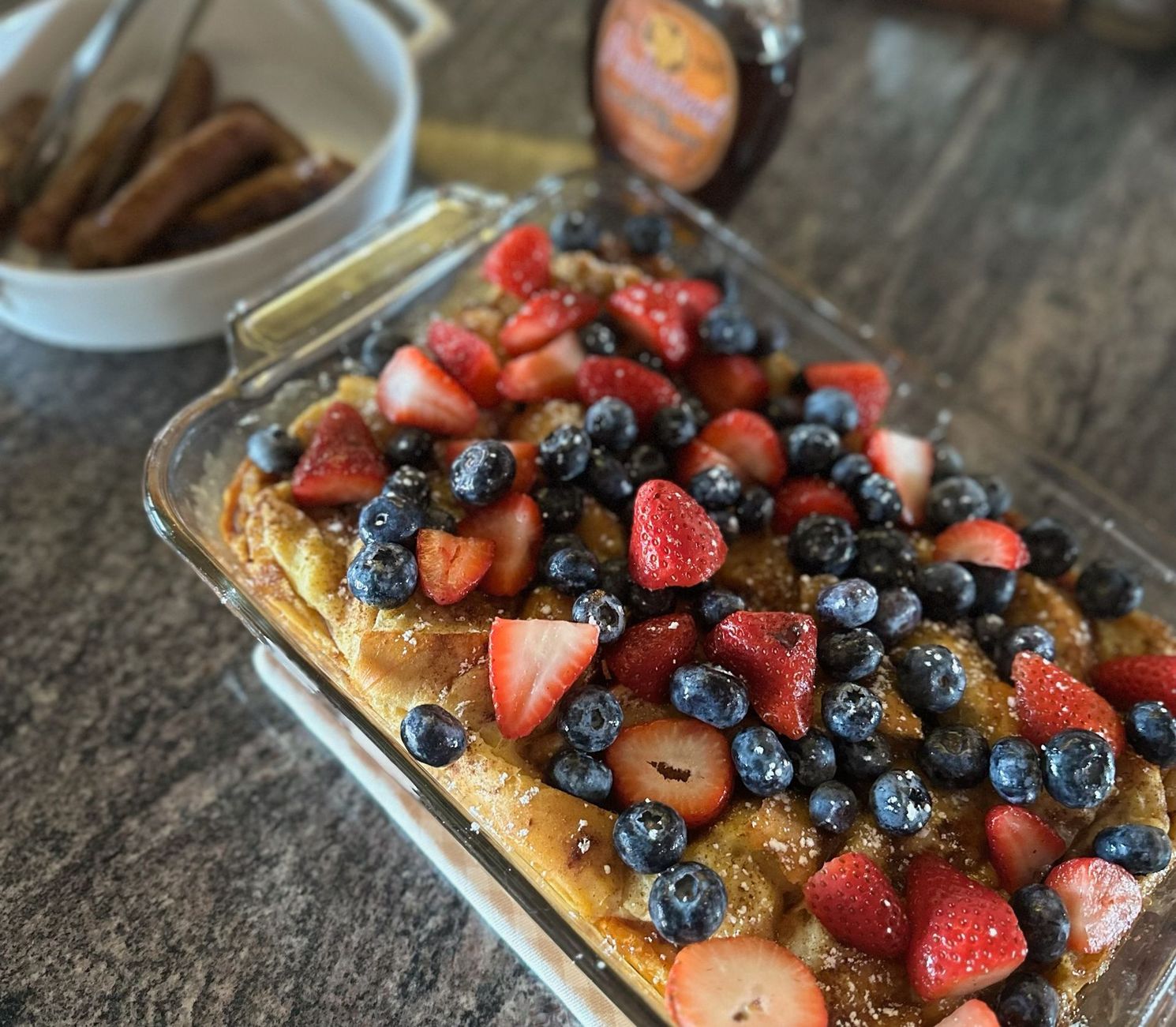 A casserole dish filled with strawberries and blueberries next to a bowl of sausages.