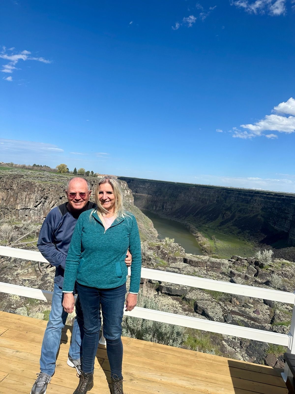 Couple poses on a wooden deck overlooking a canyon with a river. Bright blue sky.