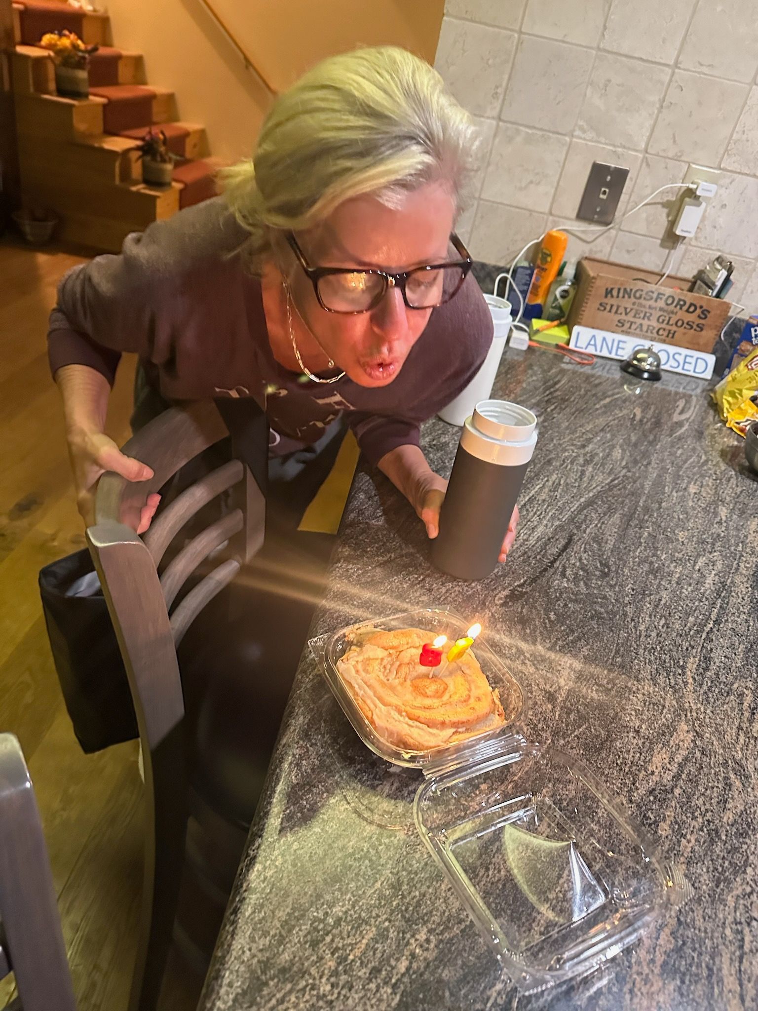 Woman blowing out a candle on a small cake in a kitchen.