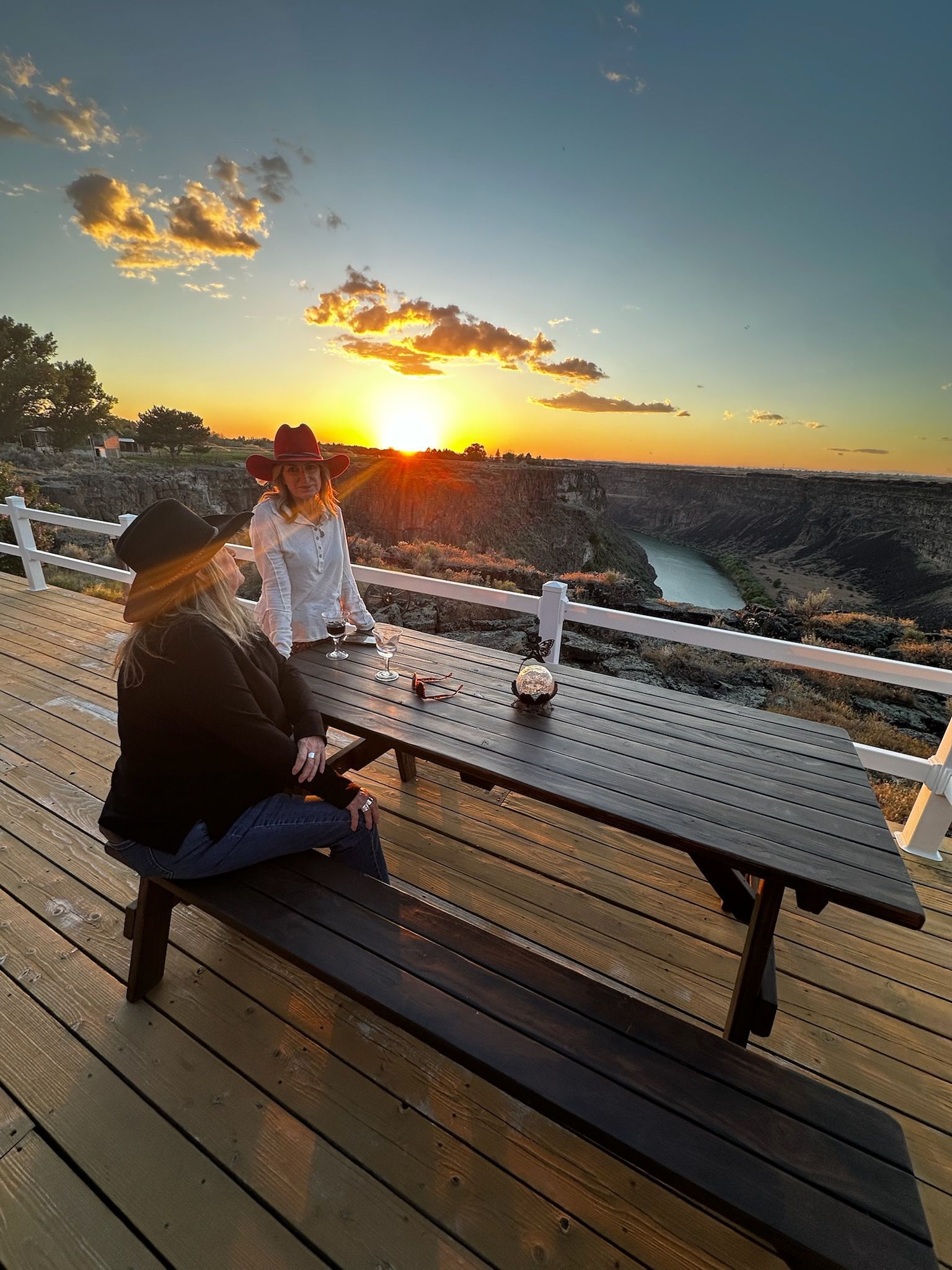 People seated at an outdoor deck table watching a sunset over the water.