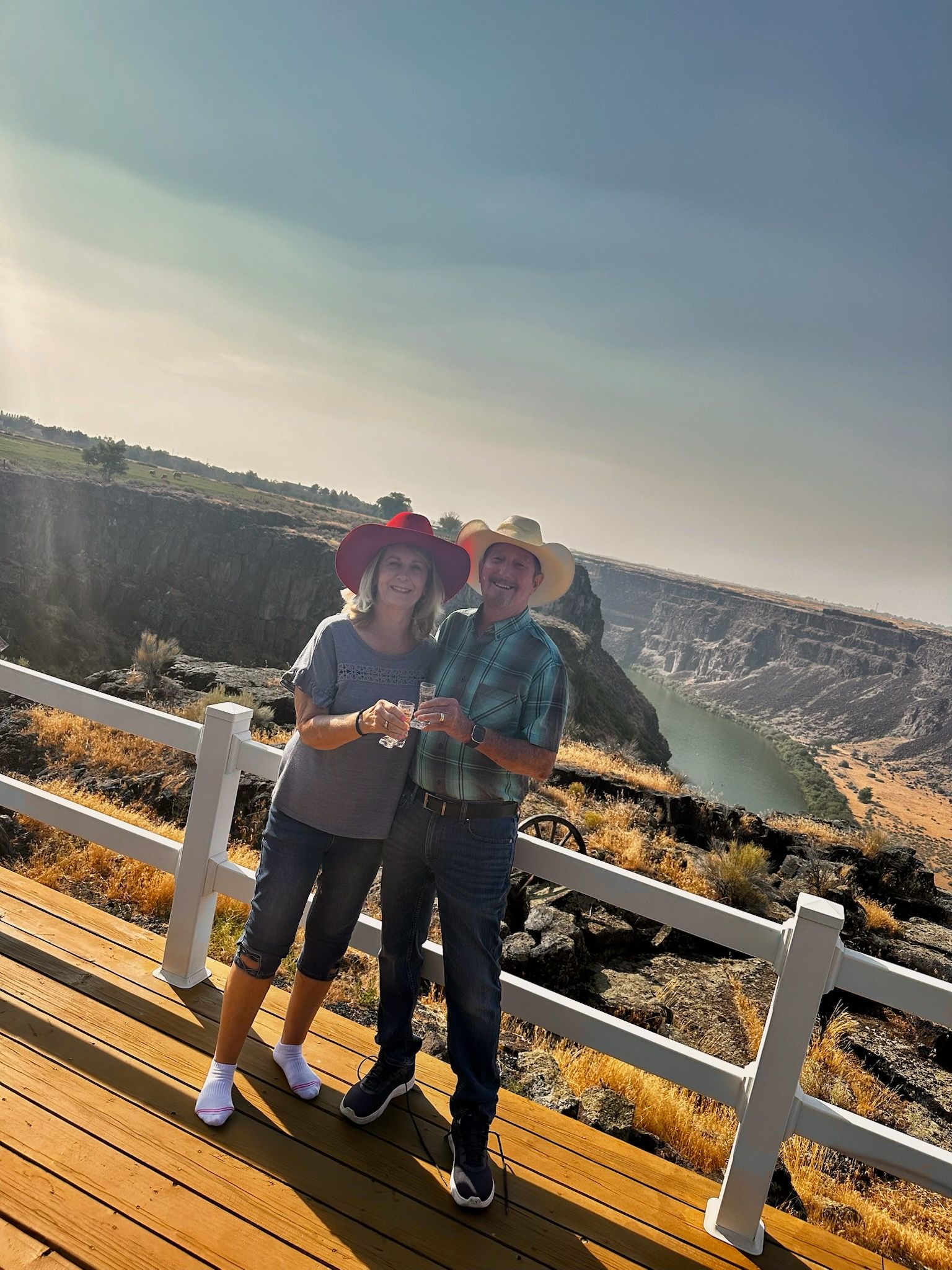 Two people in hats posing by a white fence at a scenic overlook with a lake and hills behind them.
