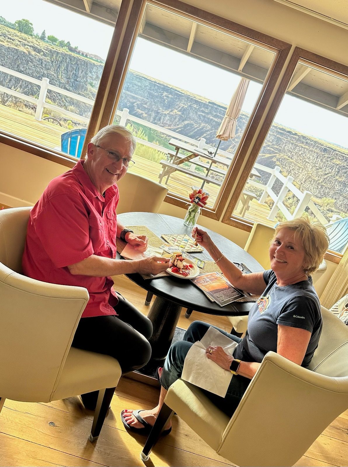 Couple at a table, smiling and eating, with a scenic view in the background.