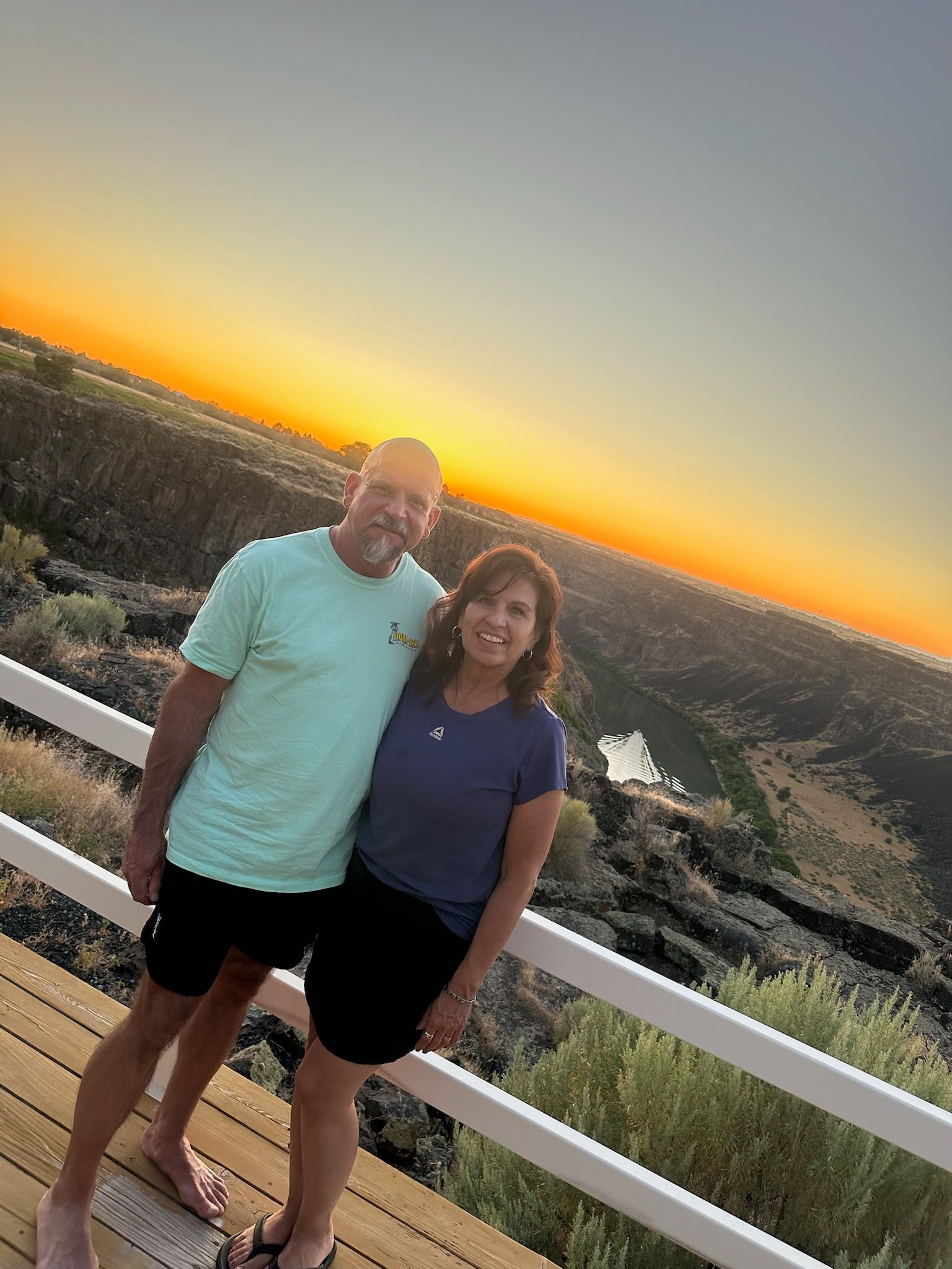 Couple poses on wooden deck at sunset. Man in turquoise shirt, woman in blue, overlooking canyon.