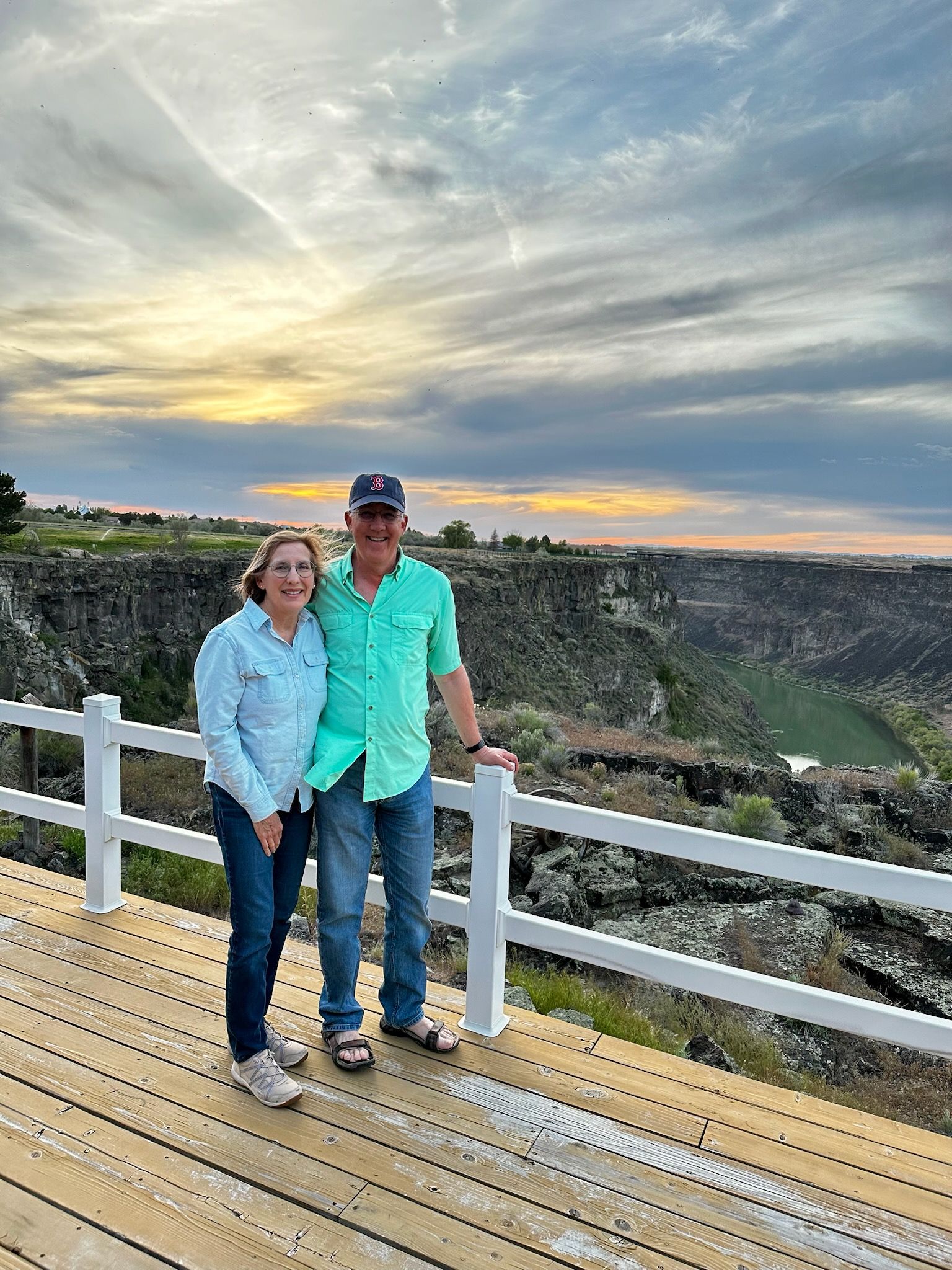 Two people standing on a white railing deck at sunset, with a sweeping canyon view behind them.