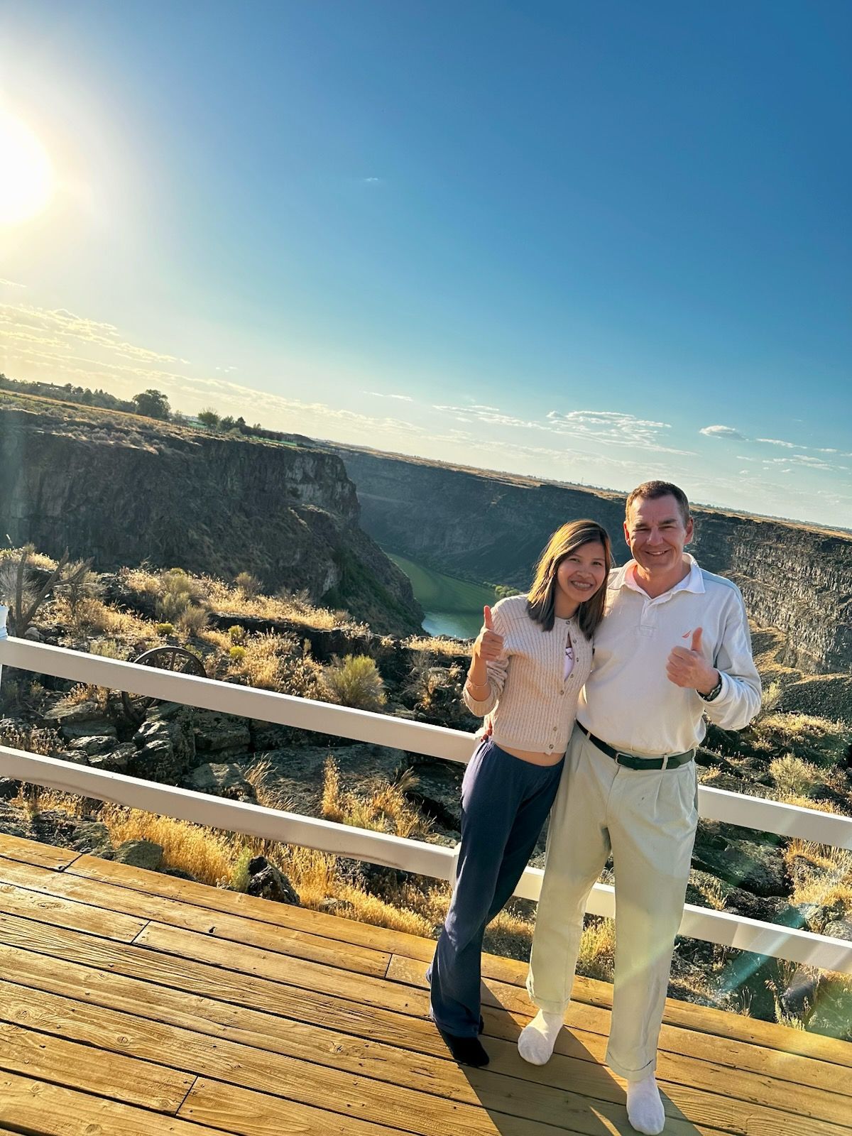 Two people smiling on a wooden deck overlooking a canyon under a bright blue sky.