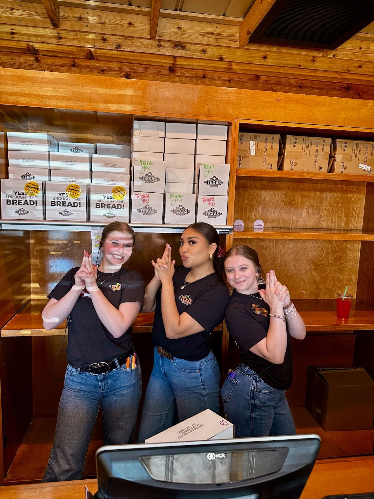 Three smiling people posing with finger guns behind a bar counter in a rustic wood-paneled room