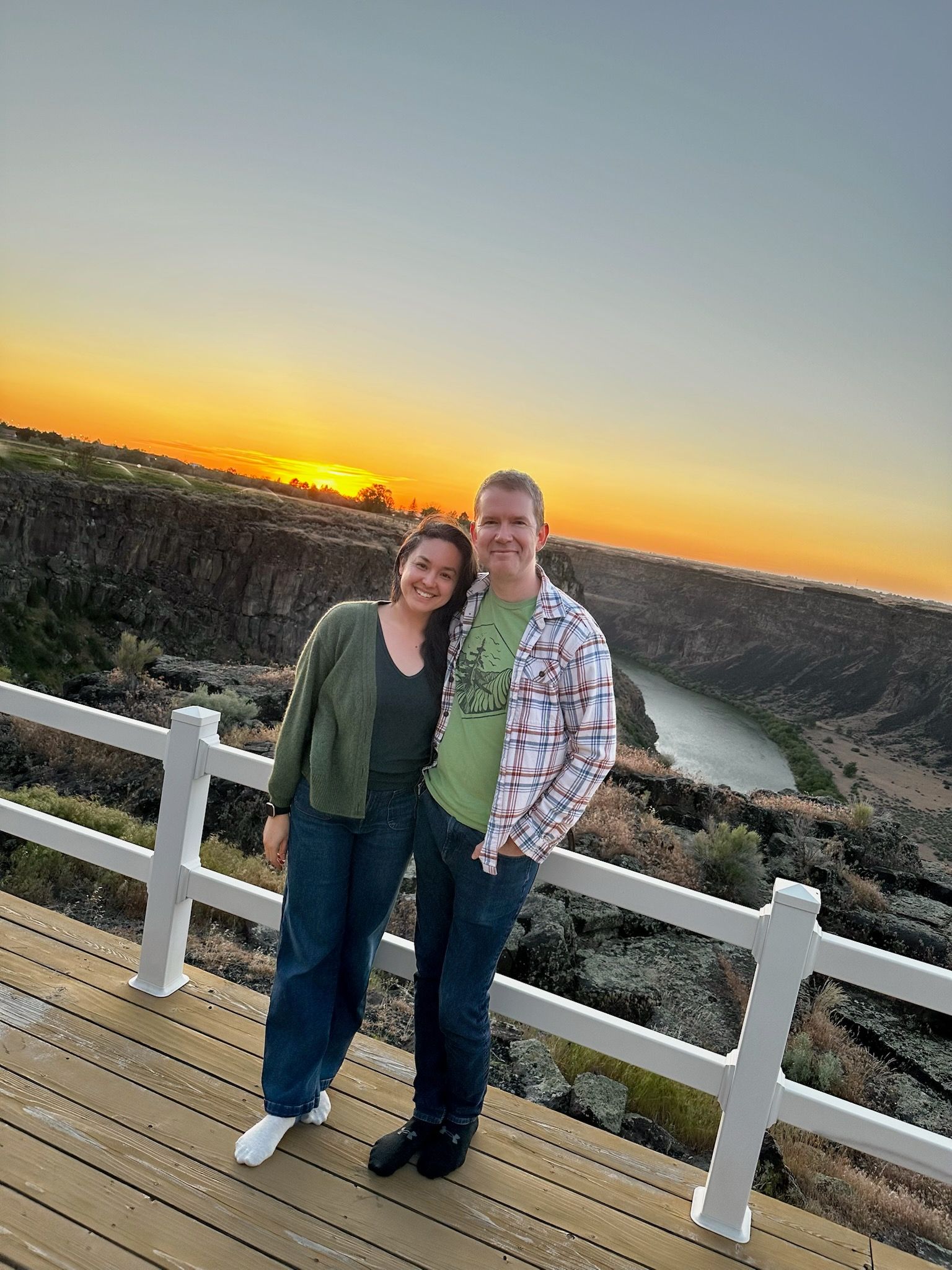Two people smiling on a wooden overlook at sunset, with a river and forested hills in the background.