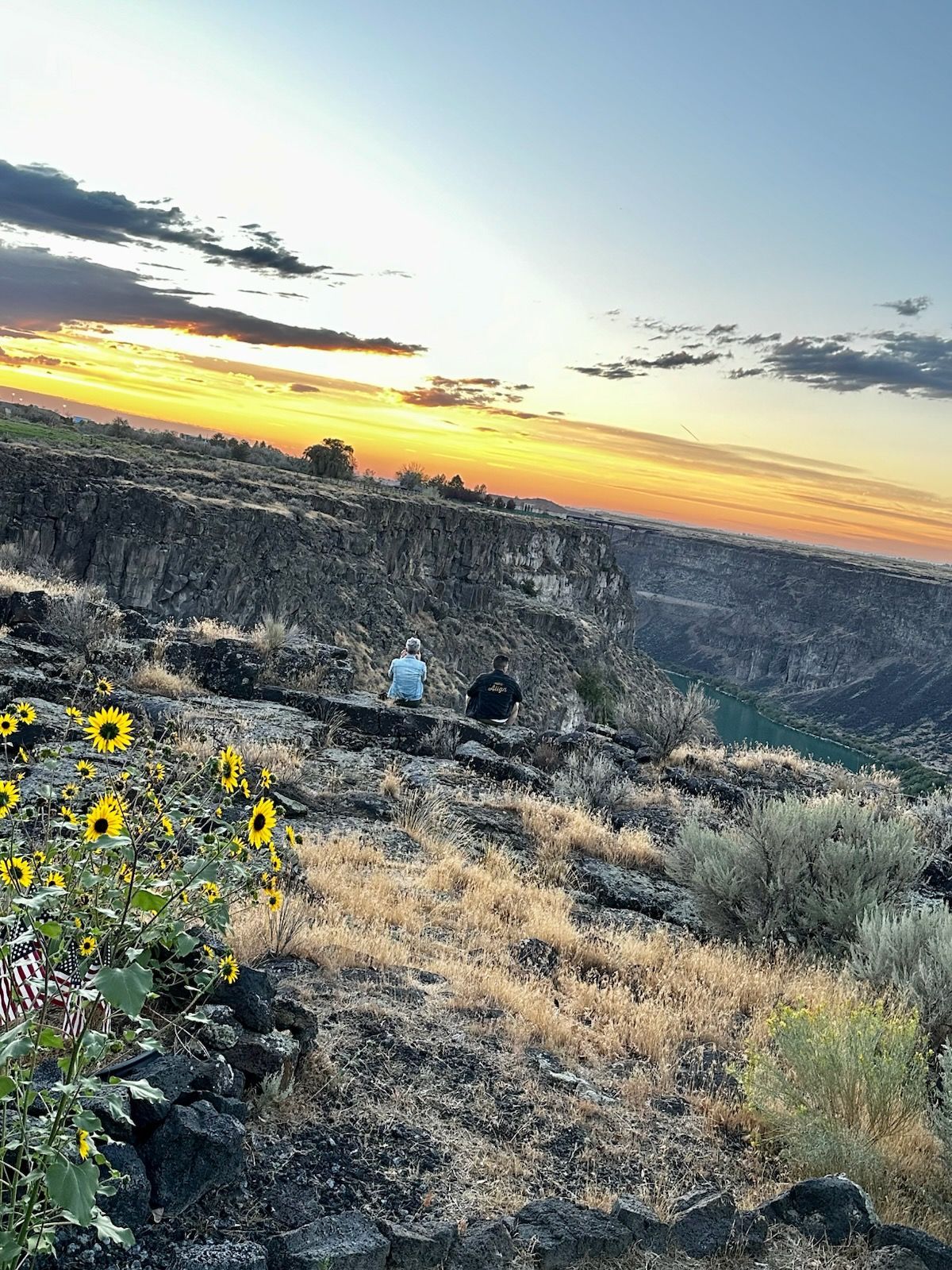 Sunset over a canyon with two people sitting on the rocky edge and yellow wildflowers in the foreground