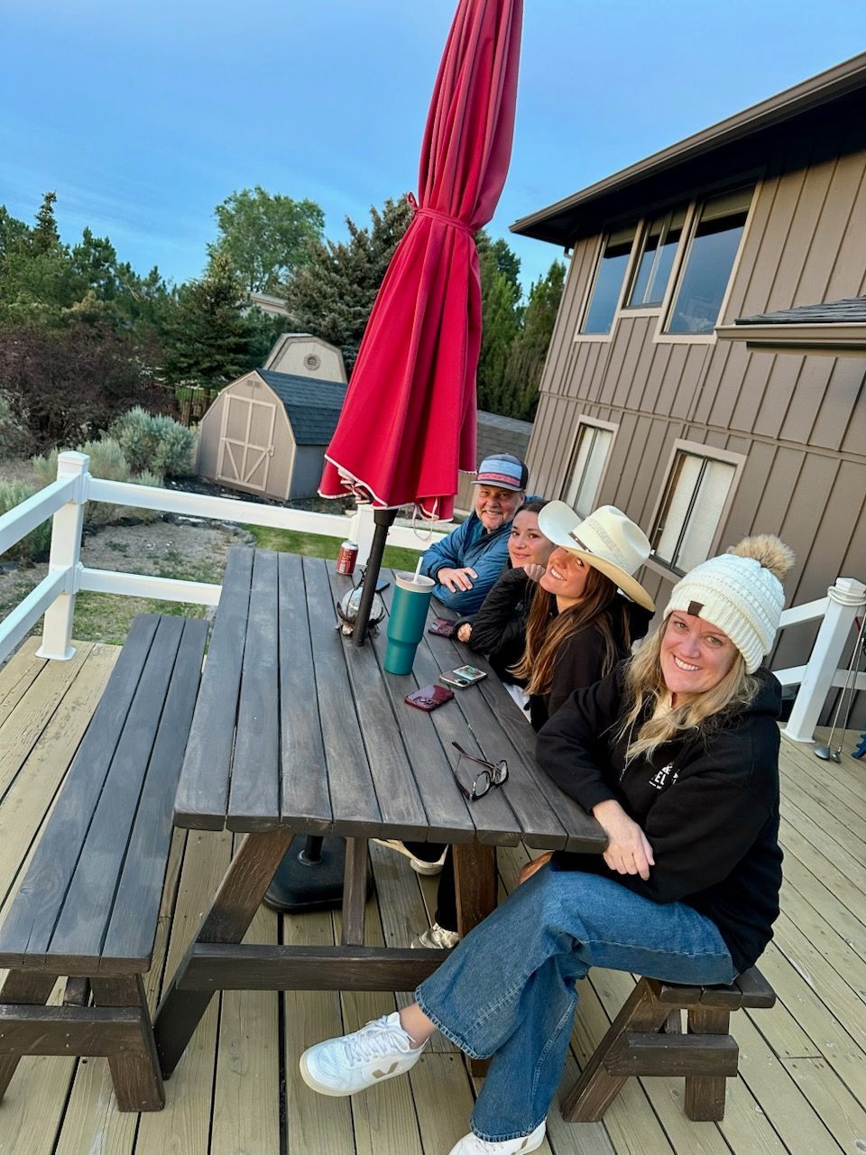 Four people sitting at a picnic table on a deck, with a red umbrella overhead.