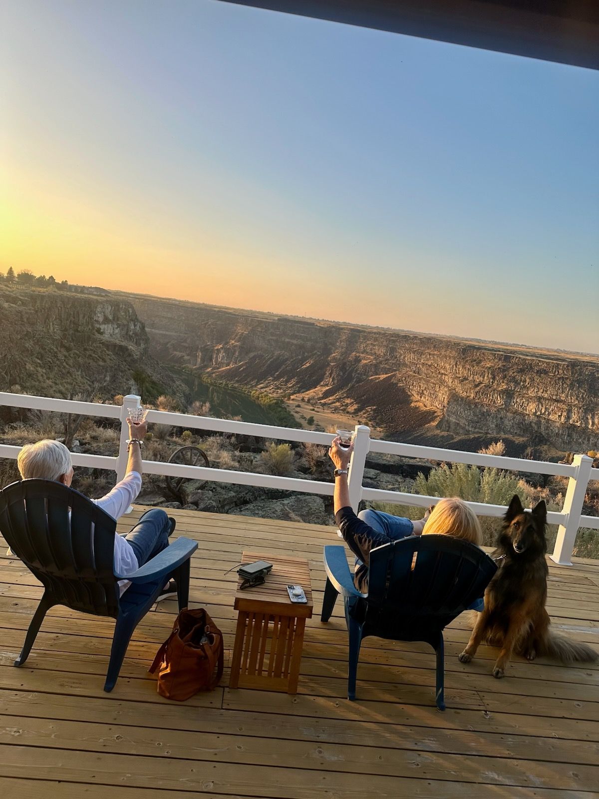 Two people relax in chairs on a deck, dog nearby, overlooking a canyon at sunset.
