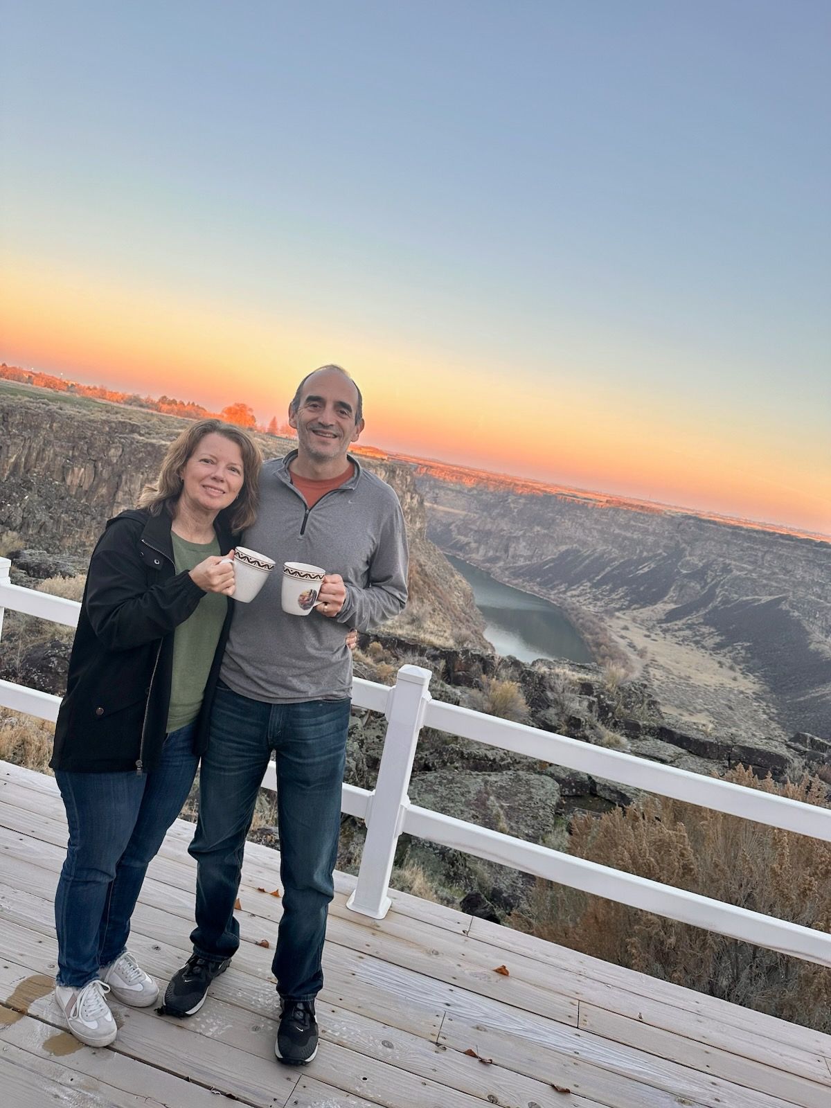 Couple on a deck, holding mugs, overlooking a canyon at sunset.