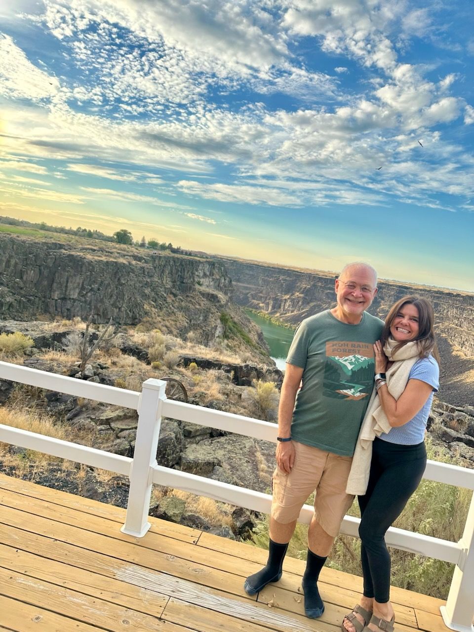 A man and woman smile on a wooden deck overlooking a canyon with a cloudy sky.