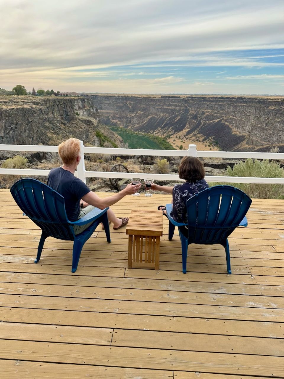 Two people in blue chairs on a deck overlooking a canyon. Cloudy sky.
