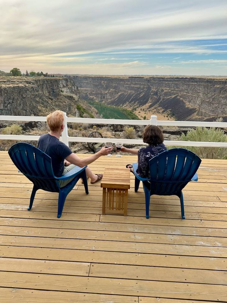 Two people sit on blue chairs on a deck, facing a wide canyon with a cloudy sky.