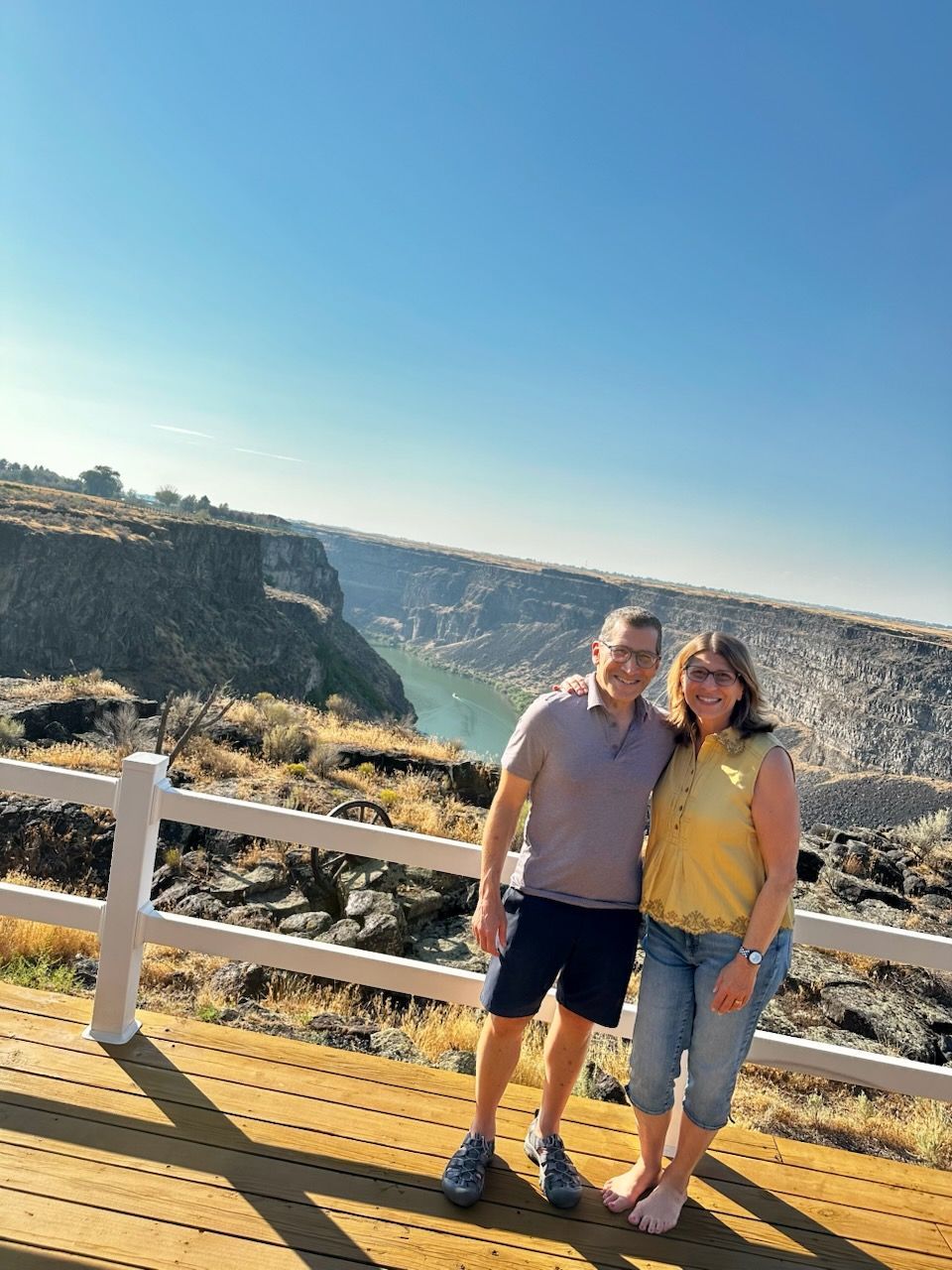 Two people standing on a boardwalk overlook with cliffs and blue sky behind them