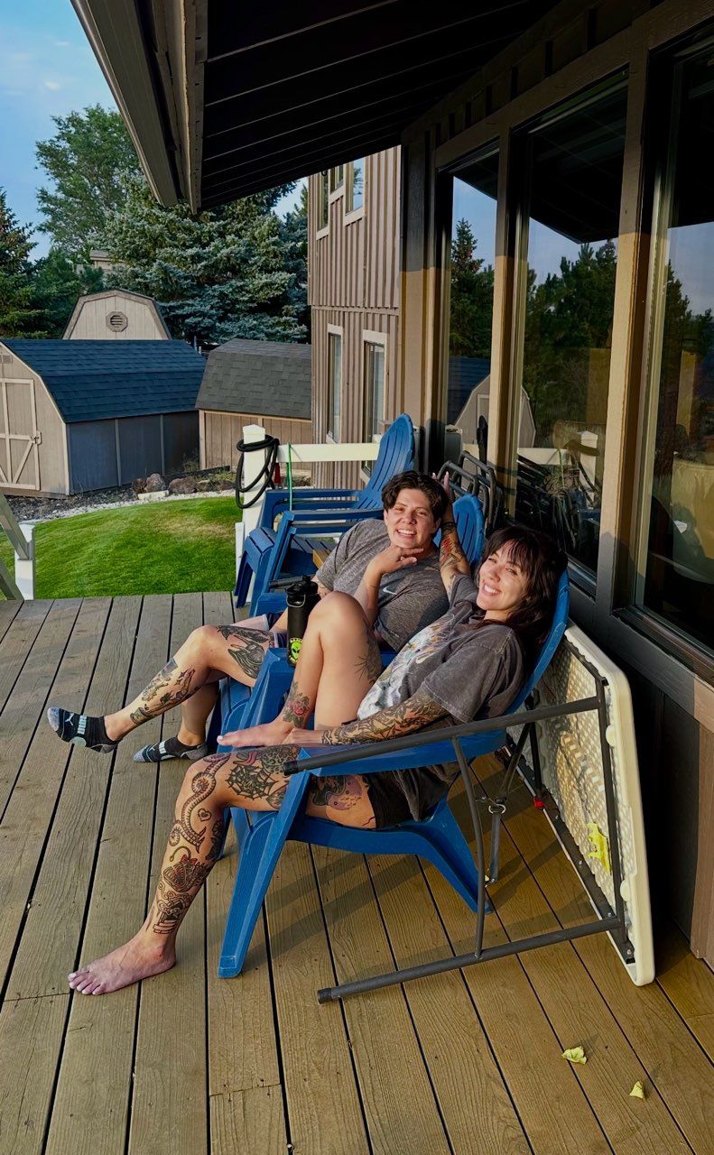 Three people relaxing in blue chairs on a wooden deck beside a glass-walled house