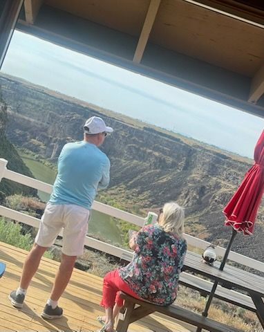 Two people on a deck overlook a canyon. Man in white hat and blue shirt, woman in floral top and red pants.
