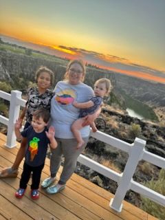 Woman holds a toddler as two children stand nearby, smiling at sunset over a canyon.
