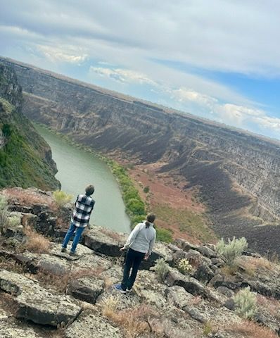 Two people stand on a rocky cliff overlooking a canyon and river under a cloudy sky.