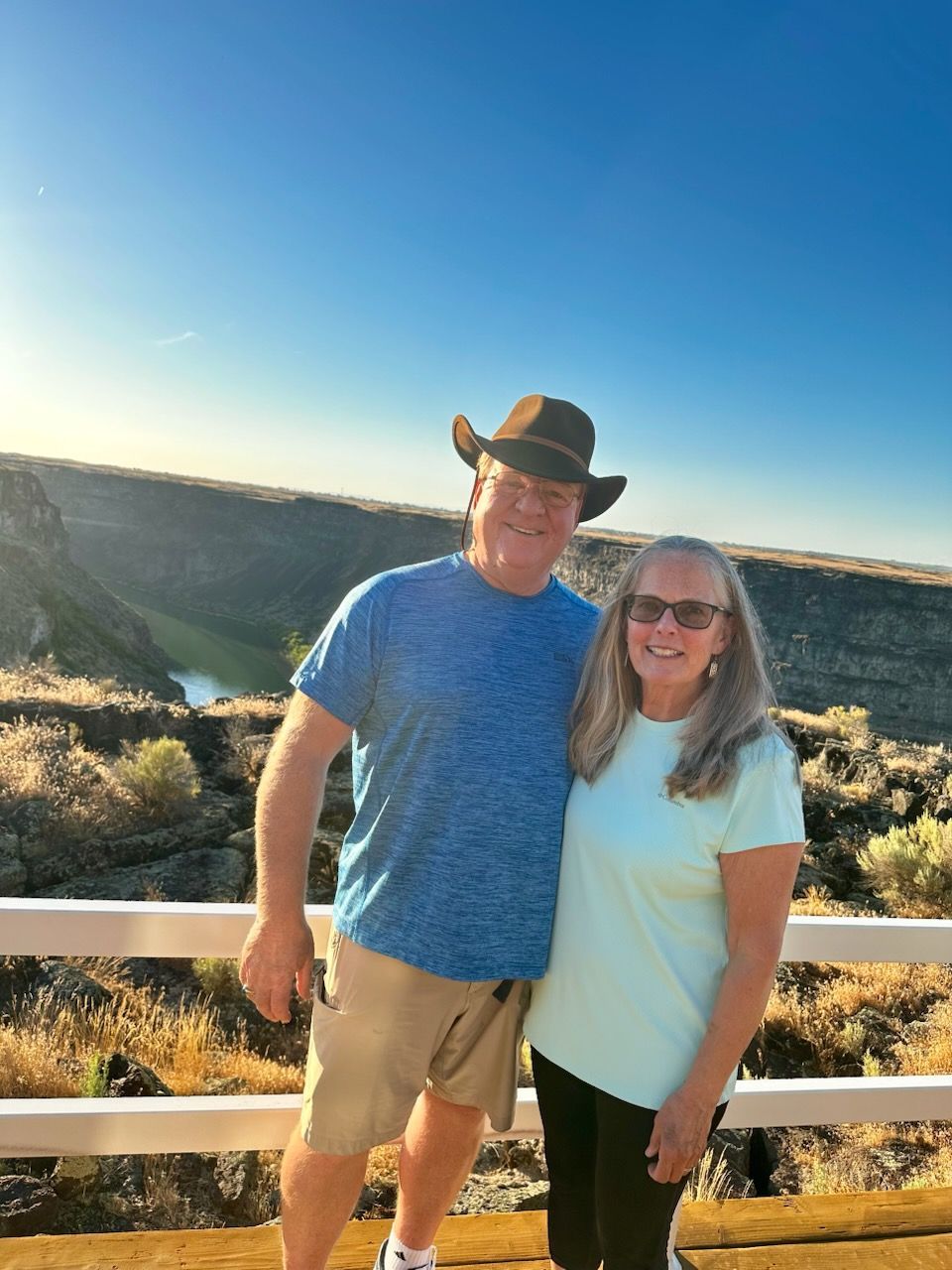 Two people smiling outdoors by a canyon railing at sunset, one in a cowboy hat.