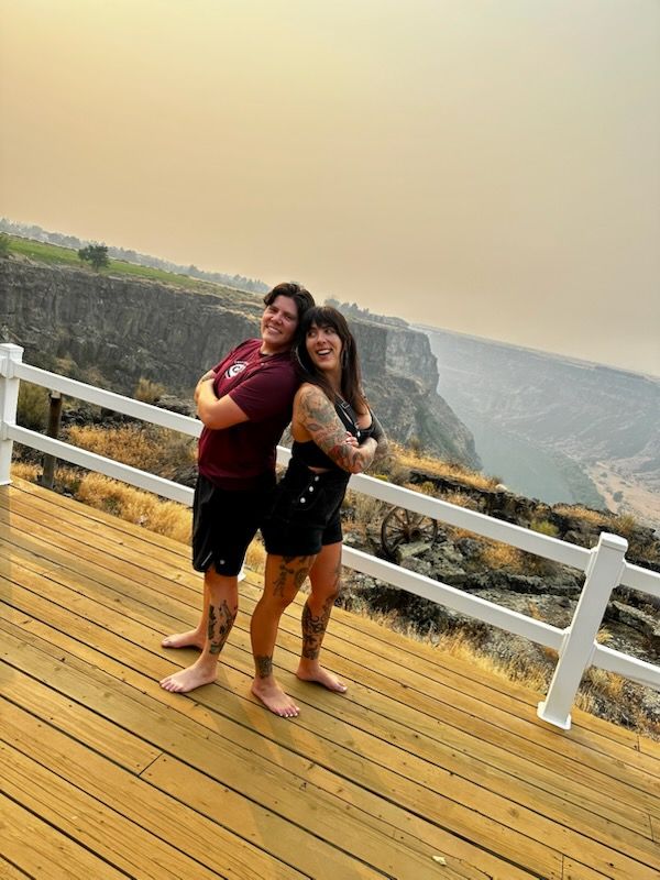 Two people posing on a wooden deck with a white railing, overlooking a hazy coastal cliff and ocean.