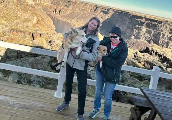 Two people holding a husky on a wooden deck with canyon cliffs in the background