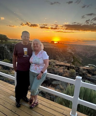 Couple on a deck at sunset overlooking a canyon. Man in maroon shirt; woman in tie-dye. Orange sky.
