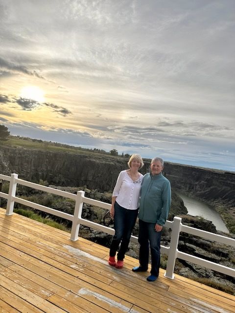 Two people standing on a wooden deck at sunset, overlooking a river and cloudy sky.