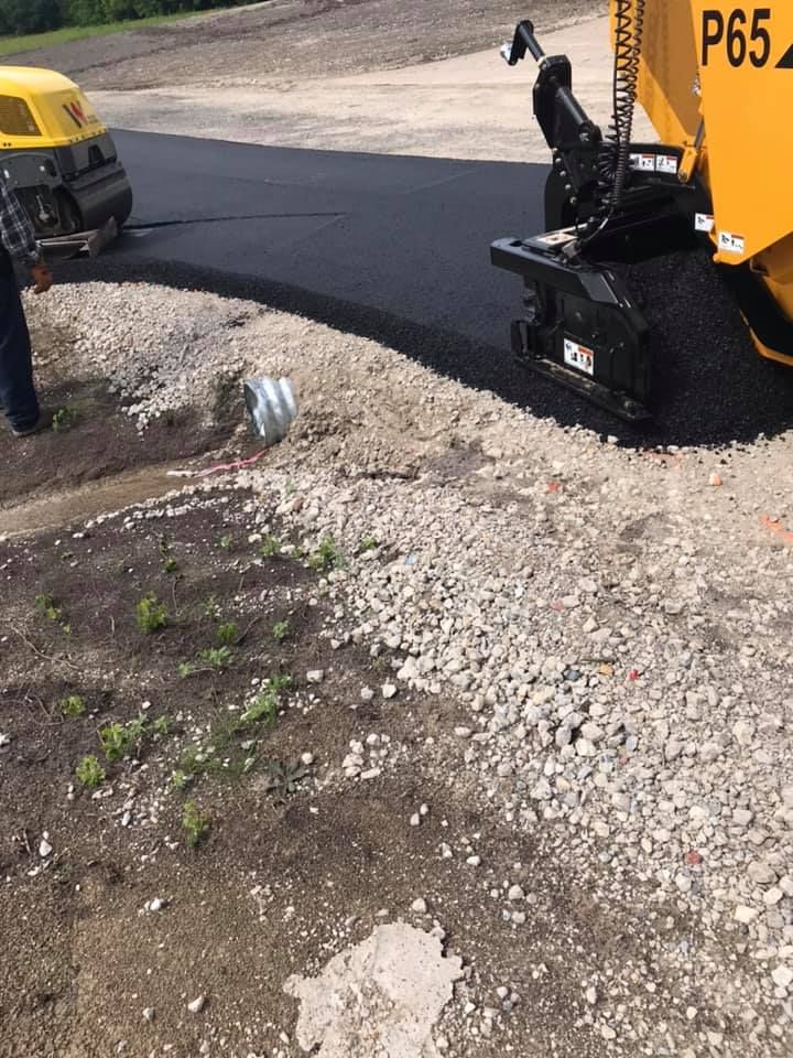A yellow truck is laying asphalt on a gravel road.