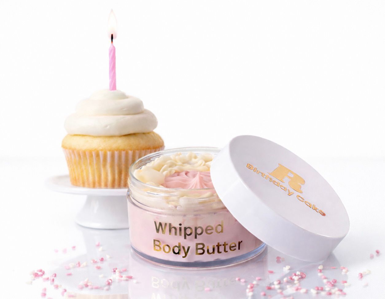 Een potje opgeklopte bodybutter naast een cupcake met een roze kaarsje op een witte achtergrond.