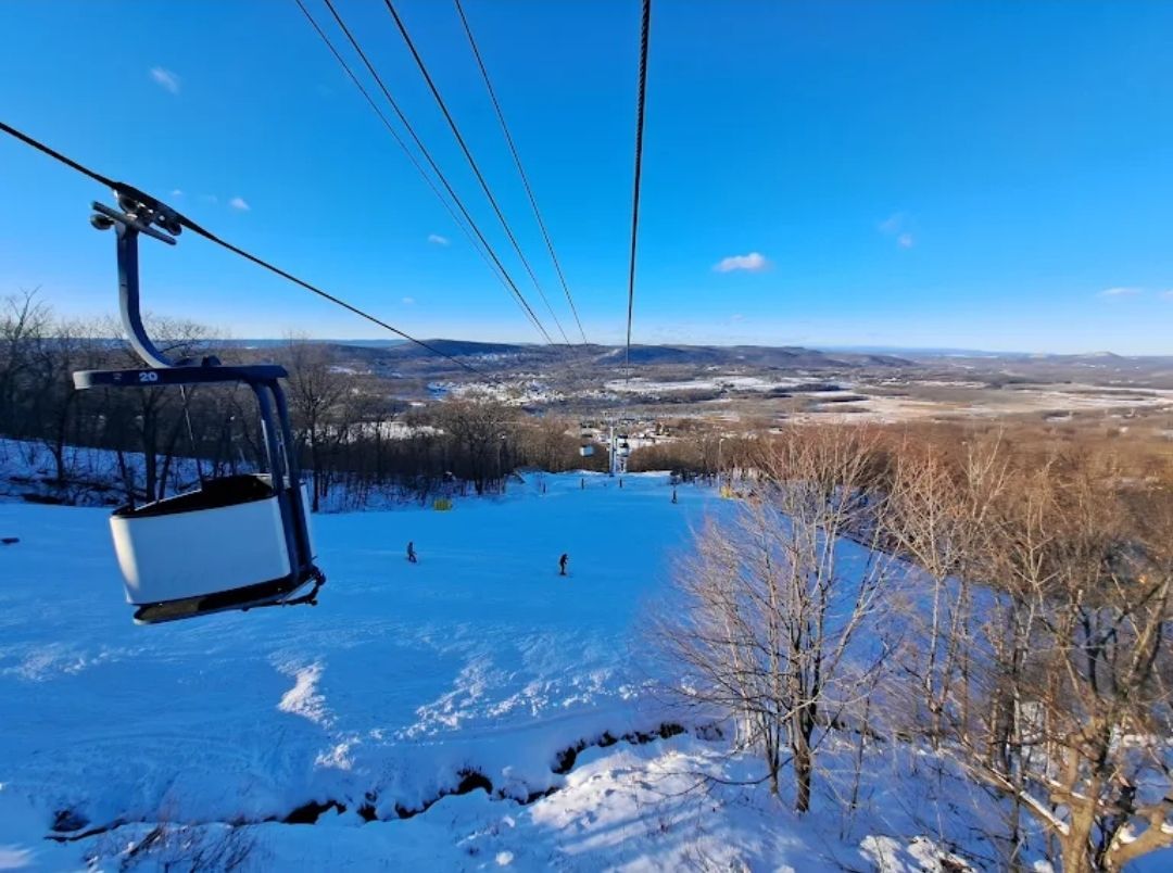 A ski lift car ascending, overlooking a snow-covered ski slope and distant landscape under a blue sky.