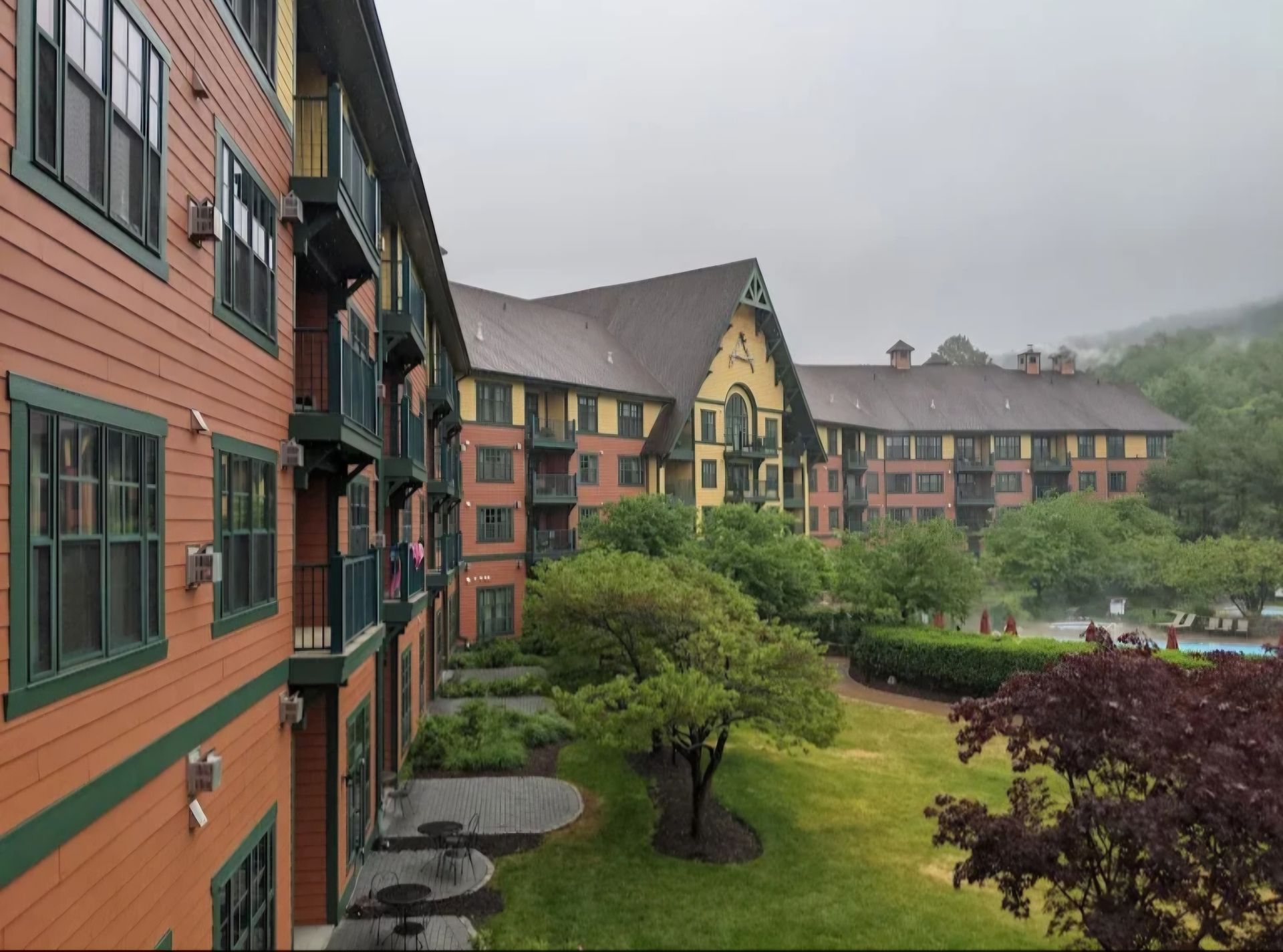 Multi-story hotel building with brown and green exterior, overlooking a grassy area and a swimming pool, on a cloudy day.