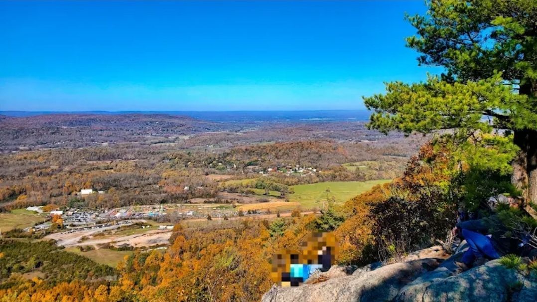Vast landscape view from a cliff edge. Overlook includes fall foliage, fields, and a bright blue sky.