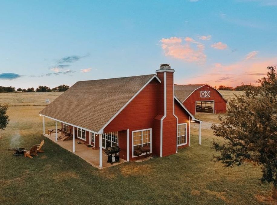 Red farmhouse and barn with a porch, chimney, and grill on a grassy field under a colorful sky.