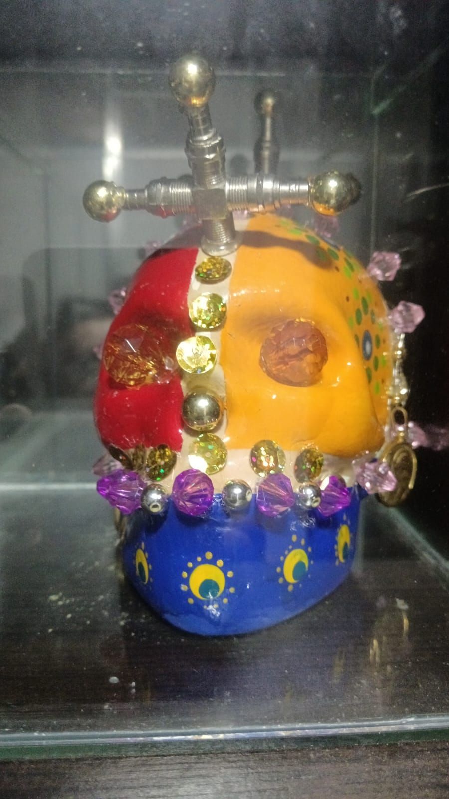 Calavera decorada: rojo, amarillo, azul y dorado. Adornada con cuentas y una cruz de metal, engastada en una vitrina.