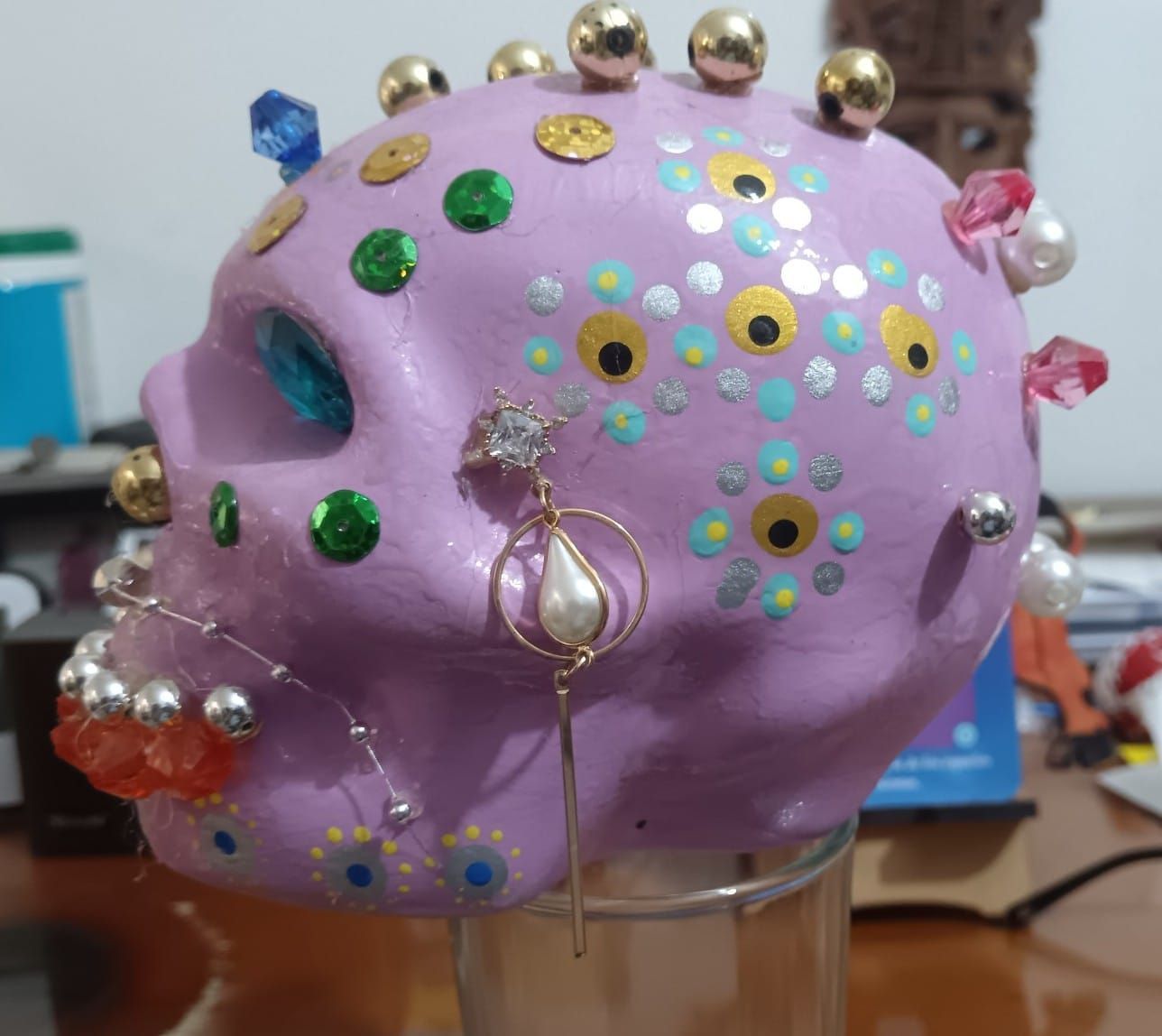 Calavera morada decorada con gemas, perlas y detalles dorados, pendientes, sobre un cristal.