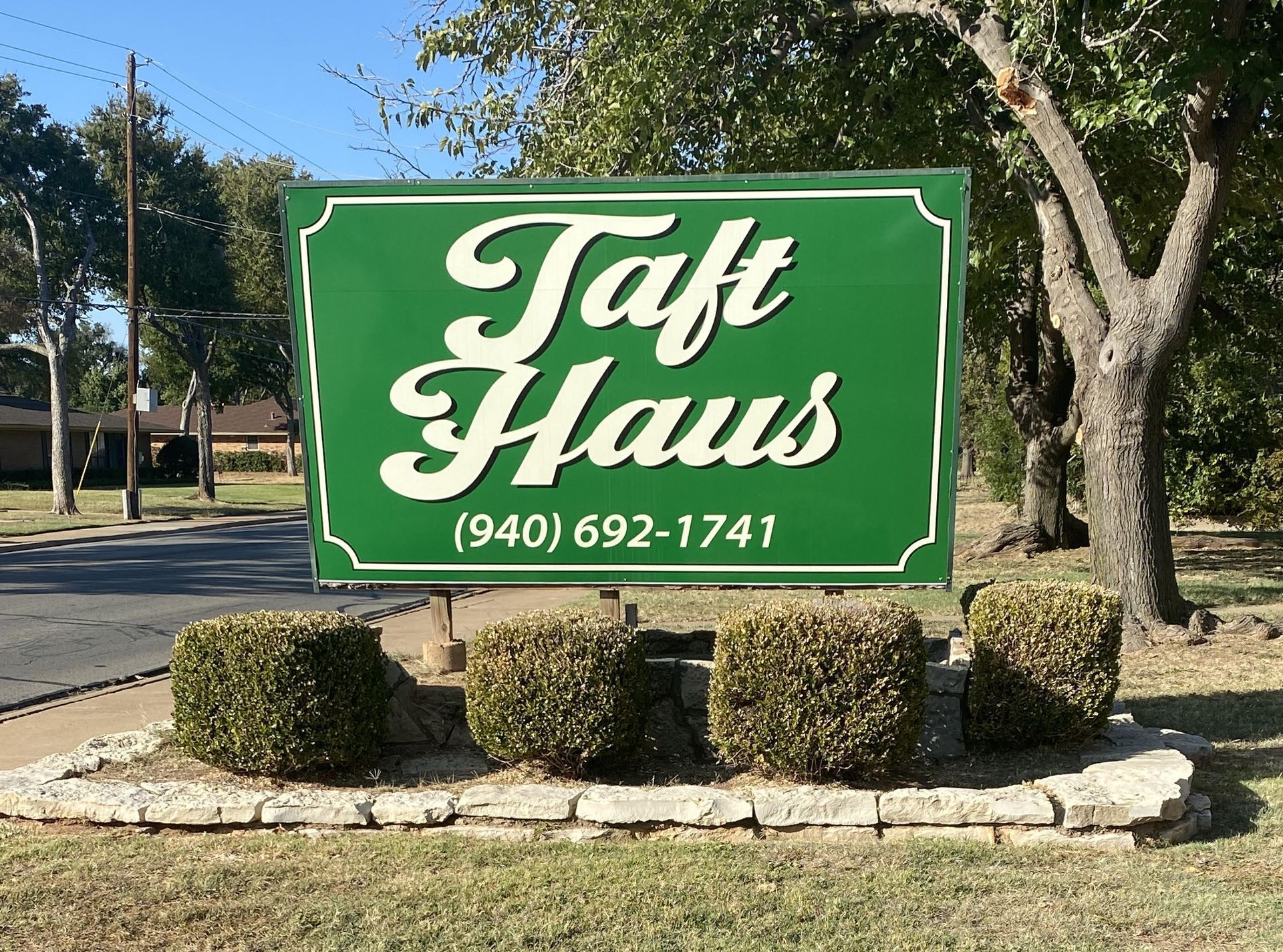 Sign for "Taft Haus" (940) 692-1741 on green background, white cursive text, trimmed bushes in front, outdoors.