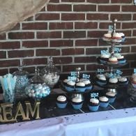 Dessert table with blue and white cupcakes, candy jars, and a tiered stand against a brick wall.