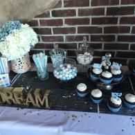 Dessert table with cupcakes, flowers, candy jars, and a gold “DREAM” sign against a brick wall