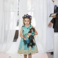 Child in a turquoise dress holding a doll, standing in front of white curtains.